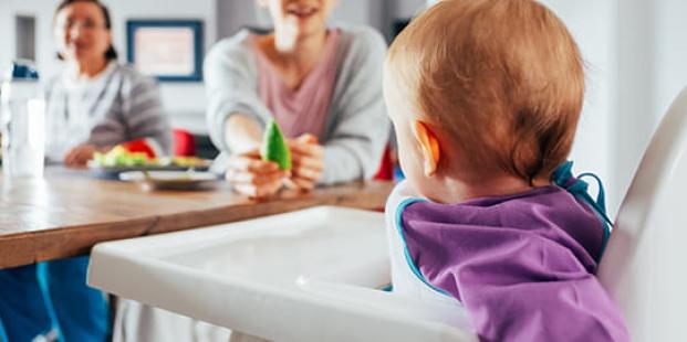 Bambino in seggiolone osserva due adulti seduti a tavola.